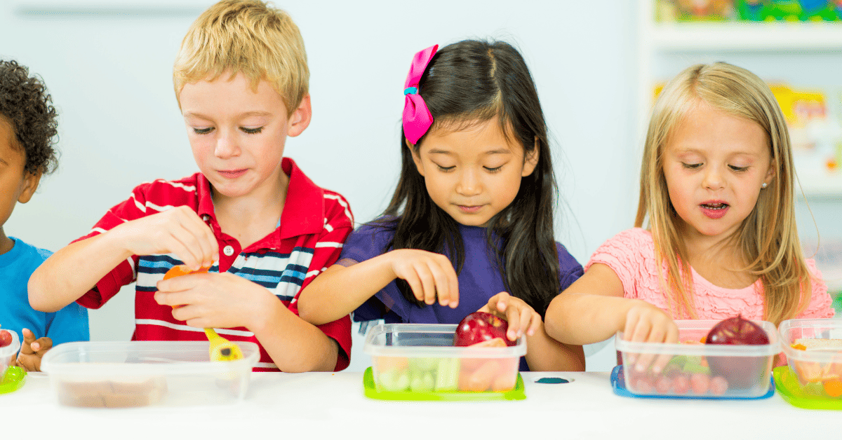 Children sitting together at school, happily eating healthy snacks from their lunchboxes, with a variety of fruits, crackers, and vegetables on the table.